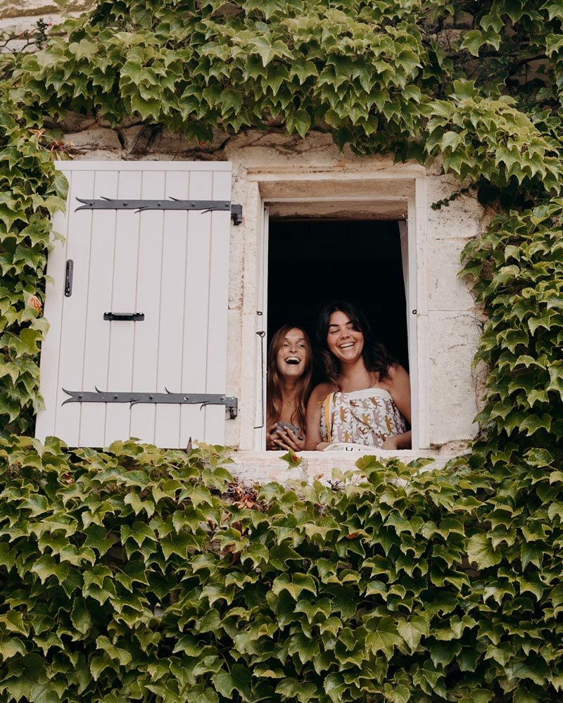 Deux invitées à la fenêtre d’un mas en pierre couvert de lierre pendant un week-end de mariage à la Bastide de Lussan près d’Uzès
