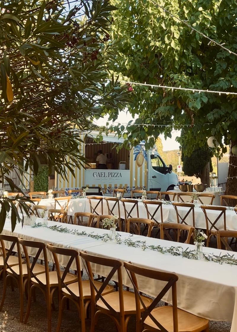 Longues tables en U  nappées de blanc installées dans la cour provençale de la Bastide de Lussan. Murier et laurier en avant plan. food truck en arrière plan.